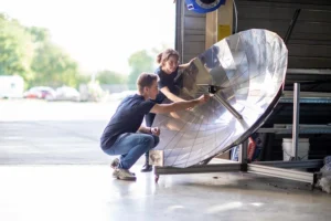 Deux étudiants ajustant un réflecteur parabolique dans un espace ouvert de type atelier, avec un environnement extérieur visible en arrière-plan.