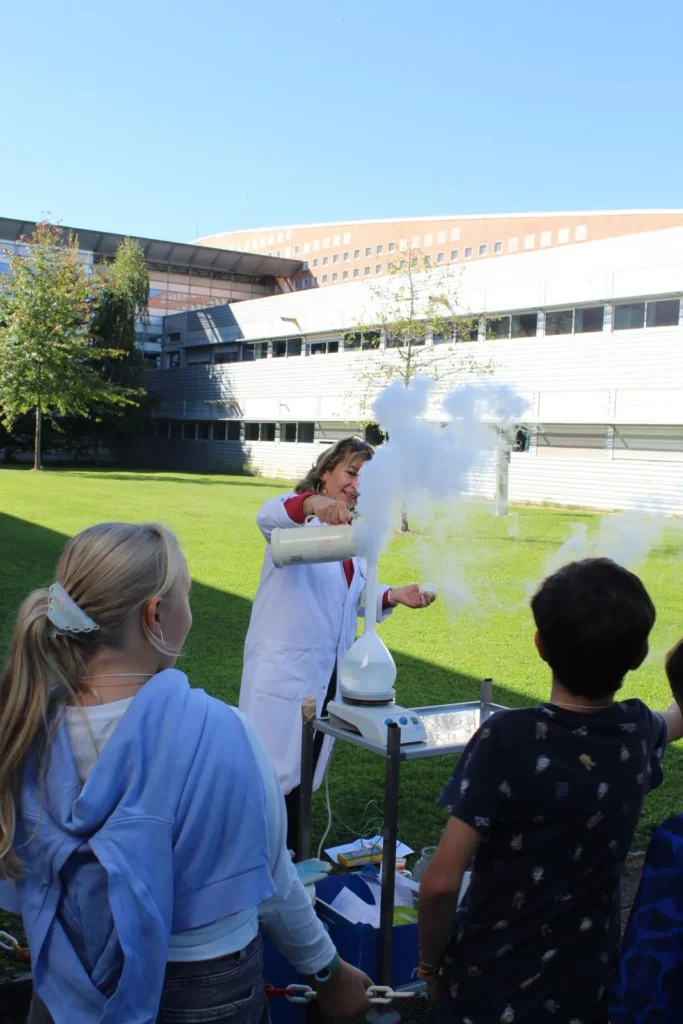 Une dame en blouse blanche qui fait une demonstration scientifique avec de la fumée blanche devant des enfants.